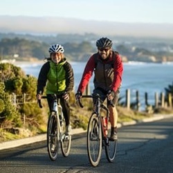 A man and a women biking along a road with a river and greenery in the background