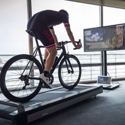 Inside of a building a person is pedaling a bike that is on a treadmill, the person is looking at a tv screen with outdoor scenery on it